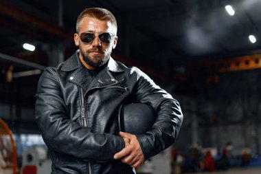 Siyah arka planda güneş gözlüklü, sakallı bir motosikletlinin portresi.