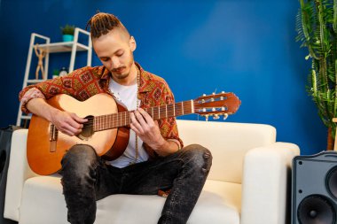 Kafkasyalı genç bir adam kanepeye oturmuş akustik gitar çalıyor.