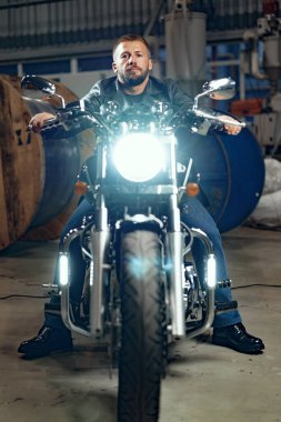 Motosikletli bir adam yeraltı otoparkında motosikletinin üstünde oturuyor.