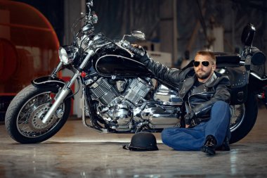 Motosikletinin yanında oturan güneş gözlüklü havalı bir motorcu.