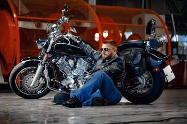 Motosikletinin yanında oturan güneş gözlüklü havalı bir motorcu.