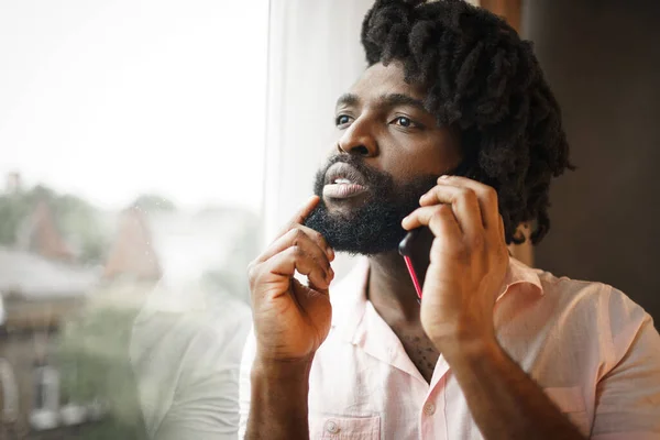 Resmi tişörtlü siyahi bir adamın pencerenin yanında telefonuyla konuşurken fotoğrafını çek. — Stok fotoğraf