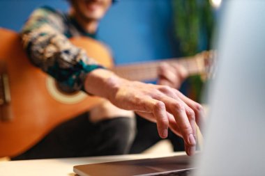 Genç beyaz adam dizüstü bilgisayarını kullanarak evde gitar dersi alıyor.