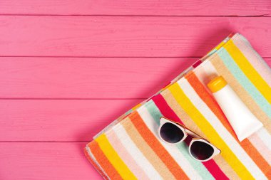 Pembe ahşap arka planda güneş kremi ve güneş gözlüklü renkli çizgili havlu.