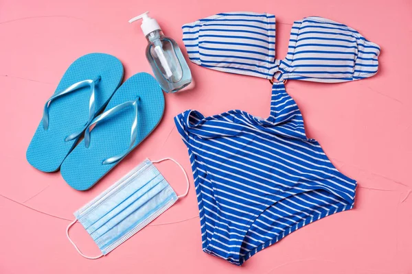 Blauer Bikini, Flip-Flops mit Schutzmaske und Händedesinfektionsmittel auf rosa Hintergrund — Stockfoto