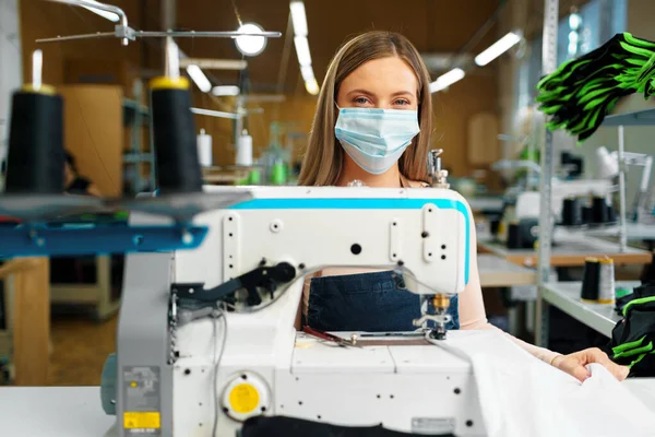 Mulher branca jovem alfaiate trabalhando na fábrica de costura usando máscara médica protetora — Fotografia de Stock