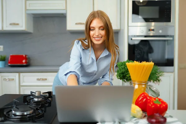 Mujer rubia joven con el pelo largo usando el ordenador portátil en una cocina —  Fotos de Stock