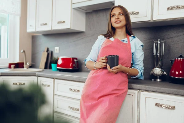 Красивая молодая женщина в красном фартуке наслаждается чашкой кофе на кухне — стоковое фото