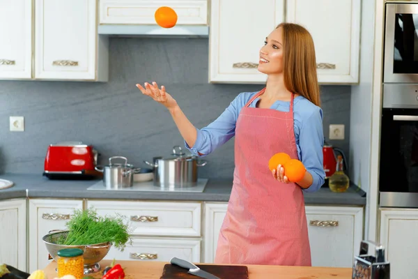 Hermosa mujer caucásica en delantal rojo sosteniendo naranjas maduras mientras está de pie en la cocina —  Fotos de Stock