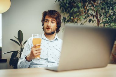 Sakallı adam bir bardak bira içerken dizüstü bilgisayarını kullanıyor.