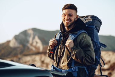Yürüyüş ekipmanlarıyla gezen genç bir adamın portresi off-road arabasının yanında duruyor.