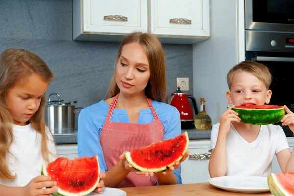 Sweet family, mother and her kids eating watermelon in their kitchen having fun