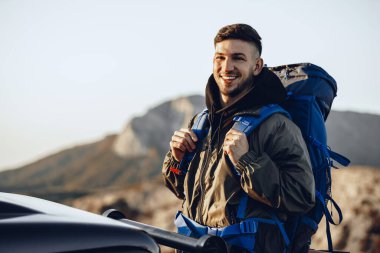 Yürüyüş ekipmanlarıyla gezen genç bir adamın portresi off-road arabasının yanında duruyor.