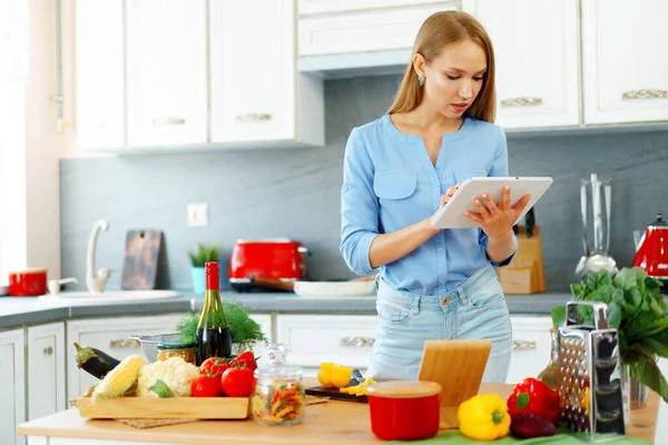 Jovem bela mulher caucasiana cozinhar e usar seu tablet digital na cozinha — Fotografia de Stock