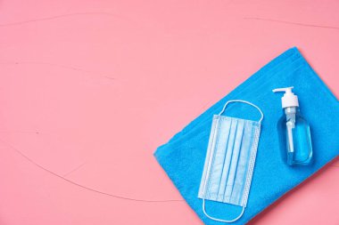 Mavi havlu, koruyucu maske ve pembe arka planda el dezenfektanı.