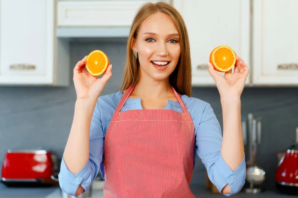 Hermosa mujer caucásica en delantal rojo sosteniendo naranjas maduras mientras está de pie en la cocina —  Fotos de Stock