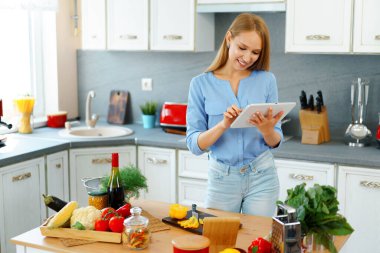 Genç, beyaz bir kadın mutfakta yemek pişiriyor ve dijital tabletini kullanıyor.