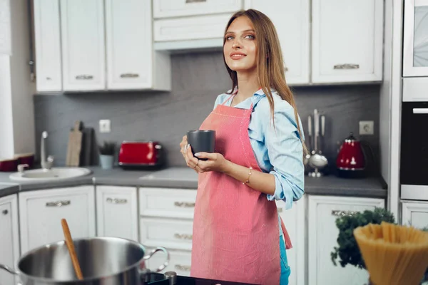 Όμορφη νεαρή γυναίκα με κόκκινη ποδιά απολαμβάνοντας ένα φλιτζάνι καφέ στην κουζίνα της — Φωτογραφία Αρχείου