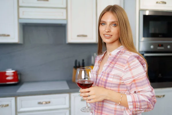 Blondynka kaukaska kobieta ze szklanką czerwonego wina stojąca w kuchni — Zdjęcie stockowe