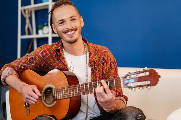 Young stylish guy musician playing guitar in his apartment