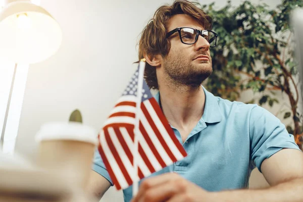 Joven hombre de negocios barbudo sentado en la mesa con portátil y bandera americana —  Fotos de Stock