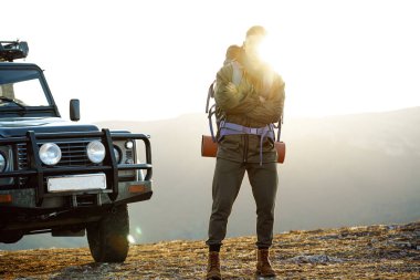Yürüyüş ekipmanlarıyla gezen genç bir adamın portresi off-road arabasının yanında duruyor.