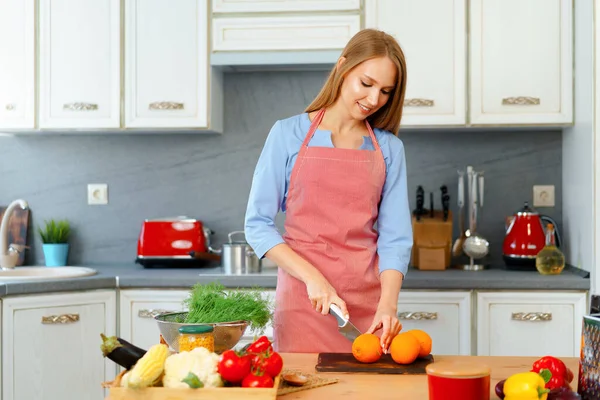 Jeune femme blonde en tablier coupant des oranges dans sa cuisine — Photo