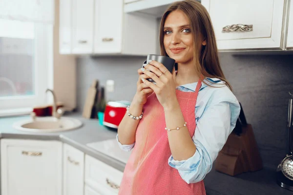 穿着红围裙的年轻漂亮的女人在厨房里喝咖啡 — 图库照片