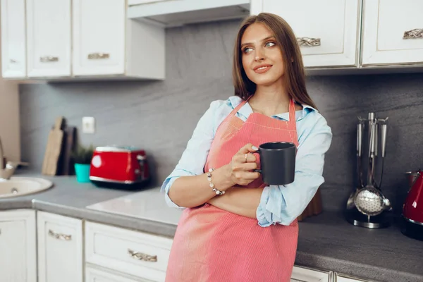 Mulher bonita em avental vermelho desfrutando de uma xícara de café em sua cozinha — Fotografia de Stock