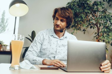 Sakallı adam bir bardak bira içerken dizüstü bilgisayarını kullanıyor.