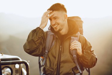 Yürüyüş ekipmanlarıyla gezen genç bir adamın portresi off-road arabasının yanında duruyor.