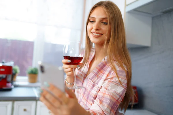Happy relaxed young woman standing in kitchen with glass of red wine and using her smartphone