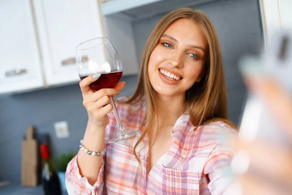 Jovem descontraída feliz em pé na cozinha com copo de vinho tinto e usando seu smartphone — Fotografia de Stock