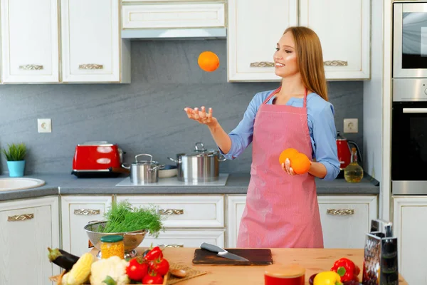 Hermosa mujer caucásica en delantal rojo sosteniendo naranjas maduras mientras está de pie en la cocina —  Fotos de Stock