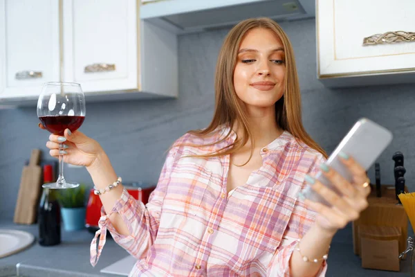 快乐而轻松的年轻女子站在厨房里，喝着红酒，用她的智能手机 — 图库照片