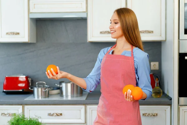 站在厨房时，穿着红色围裙的美丽的高加索女人拿着成熟的橙子 — 图库照片