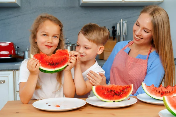 Dulce familia, madre y sus hijos comiendo sandía en su cocina divirtiéndose — Foto de Stock