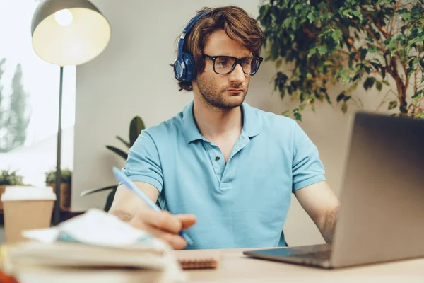 Mavi gömlekli genç adam masada oturur ve dizüstü bilgisayarla çalışır.