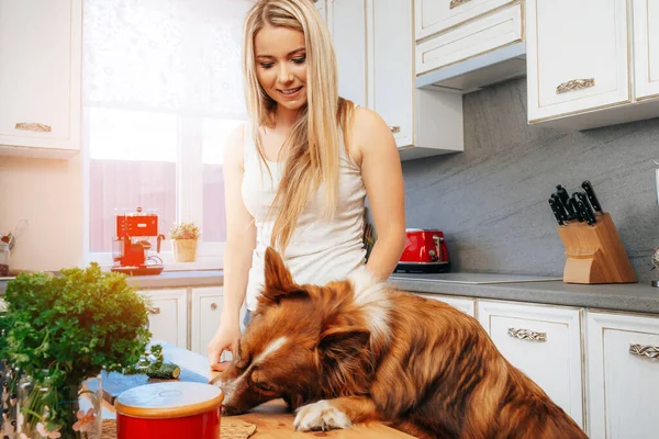 Jovem mulher sorridente preparando comida em sua cozinha com seu cão — Fotografia de Stock