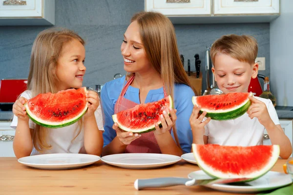 Zoet familie, moeder en haar kinderen eten watermeloen in hun keuken hebben plezier — Stockfoto