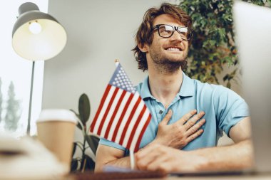 Genç sakallı iş adamı dizüstü bilgisayarı ve Amerikan bayrağıyla masada oturuyor.