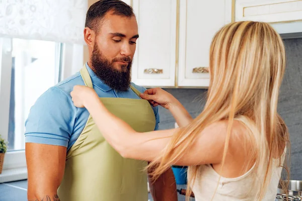 Mulher loira colocando avental em seu marido em sua cozinha — Fotografia de Stock