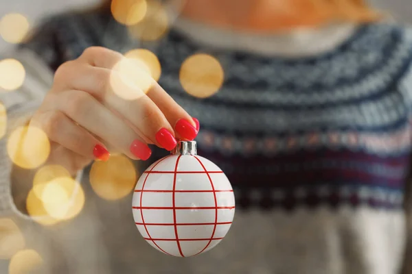 Mano di una donna in maglione festivo che tiene la bagattella natalizia con luci sfocate — Foto Stock