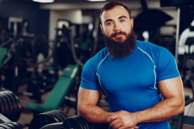 Spor salonunda duran mavi tişörtlü, sakallı, genç bir vücut geliştirici.