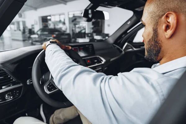 Joven afroamericano hombre sentado en un coche nuevo en sala de exposición de coches y mirando a su alrededor — Foto de Stock