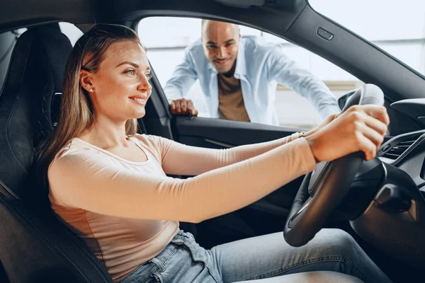 Joyful jovem casal olhando ao redor dentro de um carro novo que eles vão comprar em uma loja de carros — Fotografia de Stock