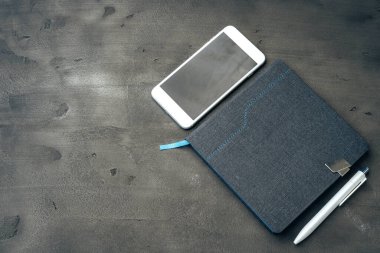 Mavi not defteri, akıllı telefon ve koyu gri masadaki kalem.