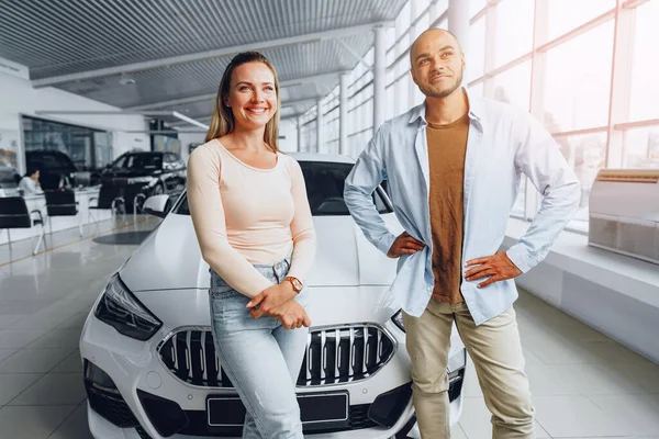 Szczęśliwa para kaukaska kobieta i afrykański amerykański mężczyzna stojący w pobliżu ich nowego samochodu wewnątrz salonu samochodowego — Zdjęcie stockowe