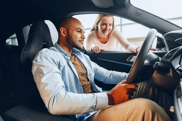 Joven pareja alegre mirando dentro de un coche nuevo que van a comprar en una tienda de coches — Foto de Stock