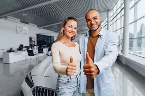 Casal feliz de mulher caucasiana e homem americano africano em pé perto de seu carro novo dentro do salão de beleza do carro — Fotografia de Stock